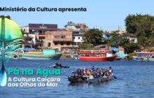 Fotografia horizontal em plano aberto. Uma grande canoa de madeira escura com jovens navega sobre água azul sob luz solar intensa. Aproximadamente 10 jovens, vestindo camisetas azuis e coletes salva-vidas, estão sentados lado a lado e remam em uníssono. A embarcação flutua no terço inferior, no centro. Em segundo plano, no rio, um homem em um caiaque preto. Ao fundo, casas e construções em alvenaria se estendem pela margem oposta. Entre elas, destacam-se: um prédio branco de 2 andares, uma construção em tijolo aparente, e um galpão com propaganda escrito "Manhattan". Três grandes barcos estão atracados na margem: um verde-água, um vermelho e um branco. A vegetação verde escura emoldura as construções nas extremidades. Céu azul claro acima. Em sobreposição à esquerda, o logo do projeto com um remo e a pena verde-azulada.