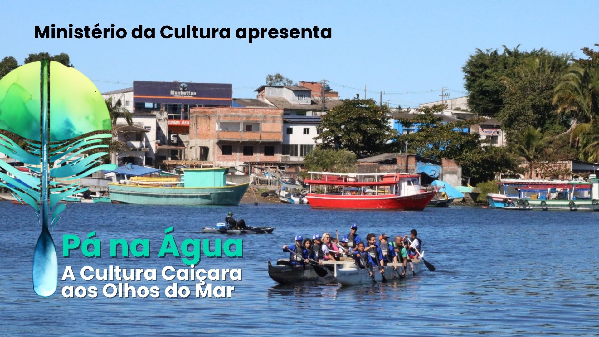 Fotografia horizontal em plano aberto. Uma grande canoa de madeira escura com jovens navega sobre água azul sob luz solar intensa. Aproximadamente 10 jovens, vestindo camisetas azuis e coletes salva-vidas, estão sentados lado a lado e remam em uníssono. A embarcação flutua no terço inferior, no centro. Em segundo plano, no rio, um homem em um caiaque preto. Ao fundo, casas e construções em alvenaria se estendem pela margem oposta. Entre elas, destacam-se: um prédio branco de 2 andares, uma construção em tijolo aparente, e um galpão com propaganda escrito "Manhattan". Três grandes barcos estão atracados na margem: um verde-água, um vermelho e um branco. A vegetação verde escura emoldura as construções nas extremidades. Céu azul claro acima. Em sobreposição à esquerda, o logo do projeto com um remo e a pena verde-azulada.