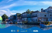 Fotografia horizontal, em plano aberto, do centro histórico de Paranaguá visto do Rio Itiberê. Em primeiro plano, a água escura do rio e, na margem direita, duas embarcações ancoradas. A primeira, à direita, é uma canoa de pesca tradicional, azul-clara e amarela, com a proa voltada para fora do quadro. A segunda é um barco de passageiros cinza, com toldo escuro. Ao longo da margem, o cais de pedra dá lugar a um conjunto de casarões históricos, com dois andares. As fachadas têm cores desgastadas: tons de azul, amarelo-claro e verde-claro. Portas e janelas são retangulares e altas. Duas palmeiras altas, de copas verdes e troncos escuros, se destacam em frente aos prédios. No centro esquerdo, uma casa menor tem telhado vermelho. No canto inferior esquerdo, uma pena estilizada verde e amarela sobre a água indica o nome do projeto: “Pá na Água”. O céu é azul claro, com nuvens brancas e esparsas. A cena combina a arquitetura colonial e o cotidiano ribeirinho em dia ensolarado. Logotipos institucionais aparecem no rodapé.