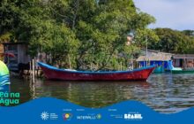 Fotografia horizontal em plano médio, focada em uma canoa de pesca tradicional ancorada. Em primeiro plano, a canoa central é vermelha, com borda azul e popa baixa, flutuando sobre a água escura e calma. A canoa está vazia e de perfil, voltada para a direita, com reflexo invertido na água. À direita, parcialmente visível, uma segunda canoa verde-escura está atracada, com uma lona azul no interior. No canto inferior esquerdo, uma pena estilizada e a inscrição "Pá na Água". Em segundo plano, uma fileira de casas simples, construídas sobre ou perto da água. À esquerda, uma pequena estrutura de madeira escura com telhado aparente. No centro, sob a densa folhagem verde de árvores e arbustos, há casas com telhados azuis. A vegetação ocupa o terço superior, fornecendo uma moldura natural. O céu, visível no canto superior direito, é azul claro. A cena transmite a tranquilidade do cotidiano ribeirinho sob forte luz solar. Logotipos institucionais aparecem no rodapé.