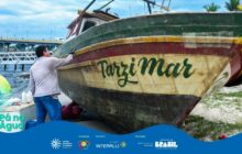 Fotografia horizontal em plano médio, com a lateral de um barco de pesca em primeiro plano. O barco está encalhado na areia, de proa voltada para a direita, e ocupa a maior parte da cena. O casco é largo e de madeira, pintado com faixas horizontais: verde-escura na parte superior, uma faixa fina vermelha e uma larga faixa amarelada abaixo, onde se lê em verde "Tarzi Mar". [PAUSA BREVE] Um homem de pele clara, vestindo calça jeans azul e camisa de manga comprida cinza-clara, está ajoelhado, com a mão esquerda apoiada na lateral do casco, olhando para o barco. A luz é difusa, indicando tempo nublado ou fim de tarde. [PAUSA BREVE] A areia é clara e molhada. No canto inferior esquerdo, aparece o logotipo do projeto "Pá na Água". Ao fundo, no terço esquerdo, uma ponte rodoviária de concreto e metal cruza a água. O local é uma orla urbana, com postes de luz e palmeiras distantes. A cena capta um momento de manutenção da embarcação. Logotipos institucionais aparecem no rodapé.