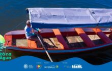 Fotografia horizontal em plano detalhe, com ângulo superior, mostrando o interior de uma pequena embarcação de madeira. O barco está no centro, em água escura e calma. A lateral do barco é laranja-avermelhada, com bordas internas e externas brancas. As travessas internas, chamadas bancos, são pintadas de um vermelho mais escuro. Na parte central, um toldo de lona cinza-clara está montado, cobrindo o remador. Um homem de pele escura e chapéu de palha sentado na popa usa uma camisa clara de mangas longas e calça escura, segurando um remo longo de madeira. Ele está olhando para a direita e se prepara para remar. A luz é forte e natural, criando reflexos intensos na superfície da água ao redor. A água preenche toda a área externa ao barco, servindo de fundo. No canto inferior esquerdo, uma pena estilizada e a inscrição "Pá na Água". A imagem celebra o trabalho e o cotidiano da vida ribeirinha. Logotipos institucionais aparecem no rodapé.