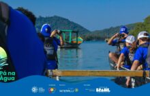 Fotografia horizontal, plano médio, vista de dentro de uma canoa. A cena mostra cinco jovens de costas, três deles em destaque, sentados e remando em uma canoa. Todos vestem coletes salva-vidas escuros e camisetas azuis, e usam bonés de cores claras, como branco ou azul. Eles não estão remando, mas sim usando câmeras fotográficas pretas para tirar fotos da paisagem. Um jovem, à esquerda, usa um boné azul e fotografa, com as costas em primeiro plano. Outros dois jovens, no centro e à direita, também estão concentrados no ato de fotografar. No centro, à frente da canoa, flutua um pequeno barco de pesca tradicional, de casco branco e toldo verde brilhante, com detalhes em amarelo e vermelho. A água, azul escura e calma, preenche o segundo plano. No horizonte, uma grande montanha coberta por mata verde-escura domina a paisagem. O céu é azul-claro, e a iluminação é suave. Na base, há uma faixa azul com o logo do projeto e os créditos de patrocínio.