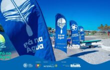 Fotografia horizontal em plano aberto. A cena se passa em uma praia com areia clara, sob luz solar intensa. Em primeiro plano, estão quatro grandes bandeiras flag verticais de tecido azul. A primeira, maior, está à esquerda e exibe o logo e a frase "Pá na Água" em letras brancas. A segunda e a terceira bandeiras, no centro, trazem o logo do projeto com as palavras "Pá na Água", "Centro Cultural" e as hashtags, confirmando a identidade visual. A quarta bandeira, à direita, é parcialmente visível. Remos grandes, pretos e brancos, estão encostados às bandeiras. Atrás das bandeiras, há duas canoas brancas de fibra de vidro. Pessoas estão em pé na areia e na água rasa, com roupas claras e escuras, preparando-se para a remada ou para as oficinas. O fundo é composto pela água calma do rio ou baía e uma fileira de palmeiras verdes, sob um céu azul vibrante. À direita, uma ponte ou passarela se estende sobre a água. Na base, há uma faixa azul com o logo do projeto e os créditos de patrocínio.