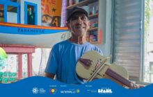 Fotografia horizontal em plano médio, tirada em ambiente interno e bem iluminado. Um senhor de pele morena e cabelos grisalhos, com um boné escuro, está em pé, sorrindo e olhando diretamente para a câmera. Ele veste uma camiseta azul-clara e tem uma expressão calorosa e orgulhosa. O senhor segura, com as duas mãos, um pequeno instrumento de cordas artesanal, semelhante a um violino ou rabeca, feito de madeira clara e marrom. O instrumento, que tem um formato delicado, está inclinado sobre seu corpo. No fundo, à esquerda, destaca-se uma maquete de barco de pesca grande, de casco branco com a palavra "PARANAGUA" em preto, e toldo azul. O interior da casa é simples, com paredes claras, prateleiras com pequenos objetos coloridos e, no canto inferior esquerdo, uma mesa rosa. A luz natural entra pela lateral direita, e a cena sugere uma oficina ou ateliê de artesanato. Na base, há uma faixa azul com o logo do projeto e os créditos de patrocínio.
