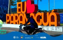 Fotografia horizontal em plano médio, com cores vibrantes. Um jovem negro, de shorts e camiseta azul com estampa branca, está sentado em uma bicicleta de cor escura, pedalando suavemente para a direita. O jovem, que usa mochila nas costas, tem a cabeça levemente abaixada e ocupa o primeiro plano. No centro, em destaque, está a famosa escultura turística com os dizeres "EU [Coração Vermelho] PARANAGUA". As letras "EU" e "PARANAGUA" são grandes, tridimensionais, e de cor laranja forte. A letra 'A' de Paranaguá está parcialmente cortada na lateral direita. O coração vermelho brilhante, no lugar da palavra "AMO", contrasta com o azul intenso do céu. O letreiro está apoiado em um muro de concreto. Ao redor, no plano de fundo, há coqueiros e uma montanha verde-escura no horizonte, à esquerda. A direita e no centro-esquerda, surgem muros com grafites e pichações. A luz solar é intensa. Na base, há uma faixa azul com o logo do projeto e os créditos de patrocínio.