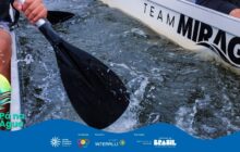 Fotografia horizontal em plano detalhe, com foco no movimento. A cena mostra a lateral de uma canoa branca, com o remo de um participante mergulhando na água. O remo, de pá preta e cabo cinza, é empunhado por uma mão visível no canto superior esquerdo. A água, escura e agitada, respinga e escorre pela borda da canoa, criando um efeito dinâmico. As ondulações e gotas de água congeladas capturam a energia da remada. Na parte superior direita da borda da canoa, as palavras "TEAM MIRAGE" estão escritas em letras pretas maiúsculas. Partes das pernas de dois remadores, vestindo calças ou shorts escuros e coletes salva-vidas com detalhes em branco e cinza, são visíveis nas laterais. A luz é difusa, mas suficiente para destacar os respingos e o reflexo na água. Na base, há uma faixa azul com o logo do projeto e os créditos de patrocínio.