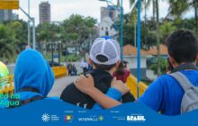 Fotografia horizontal em plano médio, tirada de costas para os personagens. A cena mostra três jovens, dois meninos e uma menina, caminhando lado a lado em uma ponte ou passarela amarela. Eles estão de costas para a câmera, com foco nítido em suas figuras. O jovem à esquerda veste um moletom azul com capuz e mochila preta. O jovem central veste uma camiseta preta e boné trucker branco e preto, com as costas em destaque. O braço do jovem da esquerda está sobre o ombro do jovem central, num gesto de amizade ou cumplicidade. O terceiro jovem, à direita, veste camiseta azul, boné e uma mochila cinza-clara, e está segurando um celular ou câmera para tirar fotos com a mão direita. A passarela, de guarda-corpo amarelo, estende-se em direção ao centro da imagem. O fundo é composto por prédios de uma cidade litorânea ou ribeirinha, incluindo uma construção antiga e branca, sob um céu levemente encoberto. Palmeiras verdes emolduram a cena na lateral. Na base, há uma faixa azul com o logo do projeto e os créditos de patrocínio.