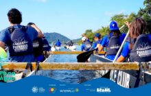 Fotografia horizontal em plano médio, tirada de dentro de uma canoa. Cerca de oito jovens, a maioria de costas, remam ativamente em uma canoa outrigger de fibra de vidro branca. O grupo está voltado para o centro e a direita da imagem, exibindo a parte de trás dos coletes salva-vidas escuros, que contêm o logo "Pá na Água Passaúna Paddle Club". Eles usam remos pretos, e a água escura e calma do rio ou baía aparece agitada pelo movimento da remada. No primeiro plano, à direita, a lateral da canoa exibe em letras pretas a palavra "PADDLE CLUB". O remador mais próximo à direita veste um boné azul virado e usa a mão esquerda para segurar o remo. O remador mais próximo à esquerda tem cabelos pretos e encaracolados e está focado na ação. A paisagem de fundo é composta por uma montanha verde e arborizada, que preenche a linha do horizonte. O céu, na parte superior, é azul-claro, com luz solar intensa vinda da esquerda, e o cenário é de atividade ao ar livre. Outras canoas com remadores são visíveis ao longe, no centro. Na base, há uma faixa azul com o logo do projeto e os créditos de patrocínio.