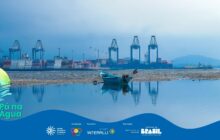 A foto horizontal, em plano geral, é visualmente dramática. Ela exibe uma vasta paisagem de maré baixa ou seca, com o porto de Paranaguá ao fundo, sob um céu encoberto. Quatro guindastes altos dominam o horizonte, ladeados por pilhas de contêineres. Em primeiro plano, uma grande poça de água espelhada reflete a cena. No centro, na areia/lama seca, um pequeno barco de pesca tradicional repousa, com a figura de uma pessoa sentada na borda. A imagem contrasta a simplicidade da vida caiçara (o barco) com a grandiosidade e a frieza da infraestrutura industrial (o porto).