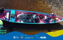 Fotografia vertical em plano geral, com uma perspectiva de cima para baixo (plano plongée). Uma canoa de madeira, longa e colorida, está ancorada em uma margem de água marrom-escura e rasa. A canoa possui o interior pintado em rosa-choque vibrante e as bordas em azul-turquesa. Cerca de cinco pessoas estão sentadas no interior do barco, a maioria voltada para a direita, misturadas à carga. A canoa está carregada com vários sacos plásticos pretos e coloridos, mochilas e cobertores, sugerindo uma viagem ou mudança. No centro, uma pessoa está agachada, vestindo um moletom cinza e um gorro. Uma outra pessoa, à direita, usa uma touca vermelha e está na extremidade da proa. O homem na popa, à esquerda, veste um casaco escuro e está em pé, olhando para a margem. A água, calma, reflete tons terrosos. A borda inferior exibe uma faixa azul do projeto e os créditos de patrocínio.