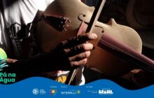 A foto horizontal, em plano detalhe, foca nas mãos de um artesão tocando um instrumento de cordas (violino ou rabeca) ainda em construção. O instrumento, feito de madeira clara e lisa (cor de bege), possui detalhes (como o espelho e o estandarte) em madeira avermelhada escura. A mão direita do artesão, visivelmente envelhecida e com a pele escura, segura o arco e fricciona as cordas. A luz do sol entra pela lateral, criando um forte contraste (chiaroscuro) que ilumina o instrumento e sombreia a mão e o rosto do artesão, conferindo um ar dramático e contemplativo. O fundo, com roupas brancas e madeiras escuras, é desfocado.