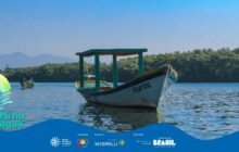 Fotografia horizontal de um rio sob céu claro, com elementos gráficos sobrepostos. No centro, uma canoa de madeira caiçara de cor clara, com o nome Kairós pintado em preto na proa. A embarcação tem uma cobertura retangular de metal verde-escura. Ela flutua em águas levemente onduladas, ocupando o centro e o terço direito da composição. Em segundo plano, à esquerda, outra canoa similar, menor, está ancorada próxima à margem. O fundo é dominado por uma densa vegetação de mata atlântica de cor verde-escura. Montanhas cobertas por neblina se estendem no horizonte esquerdo. A porção inferior da imagem é coberta por uma faixa azul curva, onde estão o logotipo do projeto "Pá na Água" à esquerda, e os logotipos de patrocinadores e realizadores, como a Lei de Incentivo à Cultura e o Governo Federal, no centro-direita. A luz natural é intensa, criando uma atmosfera calma.