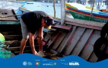 Esta peça utiliza uma fotografia de plano médio que captura um momento de trabalho manual e manutenção de uma embarcação. O forte contraste entre o homem, que está sob luz, e o interior da canoa/barco, que tem uma textura de madeira gasta, foca na ação e na cultura do trabalho braçal caiçara. O cenário de fundo, com barcos coloridos e pilares de um píer ou trapiche, complementa o ambiente portuário.