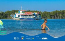 Fotografia horizontal de um estuário sob céu azul-claro, com faixa gráfica azul na base. Em primeiro plano, no canto inferior direito, um menino moreno, com aproximadamente 10 anos, está na água até a altura da cintura. Ele está sem camisa, usando apenas um calção de banho escuro, com o corpo inclinado para frente, e o braço direito estendido, apoiando-se na água calma. Sua expressão é focada. A água tem um tom azul-esverdeado intenso e pequenas ondulações. No segundo plano, centrada, uma grande embarcação de transporte de passageiros, de dois andares, está ancorada. O barco é branco e azul na parte inferior e tem detalhes vermelhos e verdes no segundo andar. Em suas laterais, diversas janelas de vidro. O fundo é dominado por uma densa faixa de vegetação verde escura, característica do manguezal. O céu é limpo e de um azul vibrante, ocupando o terço superior. A iluminação é forte e natural. A porção inferior da imagem é coberta pela faixa azul do projeto "Pá na Água", com seu logotipo e os logos de patrocinadores à direita. A cena evoca a integração da vida local com o mar.