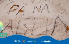 Fotografia horizontal em plano picado, focando na areia clara. A frase "PÁ NA ÁGUA" está escrita na areia, formada por galhos e pequenos pedaços de madeira. As letras "PÁ NA" na parte superior são compostas por galhos e enfeitadas com folhas secas, predominantemente amarelas e laranjas. As letras "ÁGUA" na linha inferior são feitas apenas de galhos secos e finos. Um pequeno galho na parte superior direita serve como um acento agudo. A areia, de cor bege claro, tem textura fina e parece levemente úmida, mostrando o rastro de um dedo ao lado da letra "A" de ÁGUA. A iluminação é uniforme, sem sombras fortes. Na base, há uma faixa azul ondulada com o logo do projeto à esquerda e os créditos de patrocínio no centro e na direita.