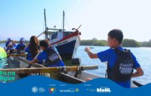 Fotografia horizontal em plano aberto, com faixa gráfica azul na base. Em primeiro plano, no interior de uma longa canoa de madeira, 4 jovens de costas ou perfil, vestindo coletes salva-vidas pretos sobre camisetas azuis, remam em águas calmas. O colete de cada um tem o logotipo "Passaúna Paddle Club". À direita, um jovem de perfil com expressão de concentração rema com o braço direito esticado. À esquerda, outros 3 jovens estão sentados, remando em direção ao fundo. O remo no canto esquerdo tem a pá verde e amarela. No centro, ligeiramente ao fundo, um barco de pesca maior, de dois mastros, branco com a proa azul e vermelha, e o nome Santa Maria pintado, está ancorado, servindo como ponto de foco da paisagem. O fundo é composto por vegetação de manguezal escura e densa nas laterais e uma porção de terra com construção à extrema direita. O céu é de um azul-claro brilhante e o sol forte. A cena transmite uma atmosfera de aprendizado e atividade física no rio. A porção inferior da imagem é coberta pela faixa azul do projeto "Pá na Água" e logotipos de parceiros.