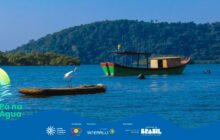 A fotografia em plano aberto capta uma cena serena do estuário, com a presença da vida selvagem (a garça) e de uma embarcação tradicional. A imagem é dominada por tons de azul e verde escuro da mata. O contraste da garça branca em primeiro plano contra a água azul e o verde da montanha cria um ponto focal forte, realçando a beleza natural da região caiçara.