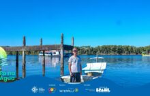 Fotografia horizontal em plano médio, sob céu azul-claro, com faixa gráfica azul na base. Em primeiro plano, no centro-direita, um homem branco de meia-idade, sorridente, com boné, camiseta cinza-clara e shorts escuros, está de pé em uma pequena embarcação de madeira branca e azul. O barco, com a proa para a direita, está ancorado na água calma. Ele ocupa o centro da composição e está emoldurado por um píer de madeira escura com pilares verticais que se erguem da água. No píer, uma pequena estrutura horizontal e escura. Em segundo plano, à esquerda, uma grande embarcação de transporte de passageiros, branca com detalhes em azul e vermelho, está ancorada. Mais ao fundo, à direita, flutua um pequeno barco de pesca amarelo e verde. O horizonte é marcado por uma densa vegetação de manguezal verde-escura. O céu é limpo e de um azul intenso, sem nuvens. A luz natural é muito forte. A porção inferior da imagem é coberta pela faixa azul do projeto "Pá na Água", com seu logotipo e os logos de patrocinadores à direita.