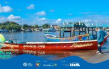 Fotografia horizontal em plano geral. Cena portuária diurna com céu azul e água calma. Em primeiro plano, uma canoa de pesca de madeira, longa e estreita, parcialmente na água e na areia escura. O casco é vermelho-vivo com faixas brancas e detalhes em azul-claro na proa e popa. Pintados na lateral, em letras brancas, os nomes "Panaguá" à esquerda e "Itauane" à direita, separados por um número de telefone e o código "CPPR". Um homem de costas, vestindo jaqueta azul-clara e calça escura, está ao lado direito da canoa, tocando-a. Em segundo plano, outra canoa de casco azul e vermelho flutua na água atrás da primeira. Ao fundo, uma fileira de embarcações atracadas e a margem com casas baixas e coloridas sob árvores. No canto superior direito, uma estrutura de píer de madeira azul. A iluminação é forte, de sol pleno. No rodapé, o logo do projeto "Pá na Água" e logotipos de patrocinadores sobre uma faixa azul escura.