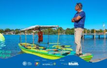 A fotografia horizontal, tirada em um dia claro, apresenta um plano médio que foca em um homem adulto e uma embarcação colorida na margem de um corpo d'água. O forte contraste entre o azul vibrante do céu e da água, o verde limão do barco e as roupas do homem cria um impacto visual imediato. A pose do homem, olhando para o horizonte, sugere contemplação ou vigilância, ligando a figura ao ambiente ribeirinho. A luz é intensa e a profundidade de campo está bem focada nos elementos centrais.