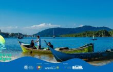 Fotografia horizontal em plano geral sob céu azul claro. No centro, três canoas de madeira tradicionais em primeiro e segundo plano flutuam em água azul-esverdeada. A canoa central e principal, amarela-esverdeada, é ocupada por três pessoas sentadas. Um jovem de camisa branca, no centro, usa um remo amarelo como apoio. À esquerda, uma jovem de cabelos longos e camisa escura. À direita, um homem de camisa escura, parcialmente oculto. À frente e à direita, a proa de uma canoa azul-escura com a frase "Obrigado Deus" pintada em branco. À esquerda, uma terceira canoa verde é parcialmente visível. Ao fundo, o rio com uma fileira de barcos a vela e barcos de pesca atracados, e um barco a vela solitário no centro. A linha do horizonte é dominada por vegetação densa e morros de tons escuros. Iluminação forte de dia ensolarado. No rodapé, faixa azul do projeto "Pá na Água" com logotipos.