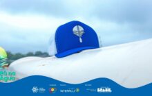 Fotografia horizontal em plano detalhe. Um boné trucker azul-marinho com tela branca traseira está posicionado no centro da imagem. O boné repousa sobre uma superfície branca, curva e larga, que atravessa horizontalmente o centro do quadro. Na frente do boné, o logotipo do projeto "Pá na Água", um remo com gotas, é estampado em branco. O fundo está ligeiramente desfocado, mostrando uma faixa de árvores e a água do rio. O céu acima é de um cinza-claro, nublado, com iluminação suave e difusa. A atmosfera é calma. A superfície branca em primeiro plano parece ser a lateral de uma embarcação. No rodapé, a faixa azul do projeto com logotipos de patrocinadores.