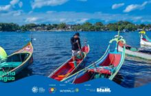 Fotografia horizontal em plano médio no Rio Itiberê. Quatro canoas de pesca tradicionais de madeira, longas e com proas elevadas e decoradas, estão flutuando em água azul-escura com pequenas ondas. Em primeiro plano, no centro, a canoa principal é vermelha internamente, com bordas externas amarelas e brancas. No centro dela, um jovem de boné preto e agasalho escuro, short estampado e tênis, está em pé. Ele segura um remo, olhando para baixo, concentrado na água. À esquerda e à direita, proas elevadas de outras canoas decoradas se projetam na composição, em cores vermelho, verde, amarelo e branco. Ao fundo, casas e vegetação densa ao longo da margem, sob um céu azul-claro com nuvens brancas. Outras embarcações estão atracadas. A luz é solar, forte e direta, criando brilhos na superfície da água. A cena representa o manejo das canoas no cotidiano ribeirinho.