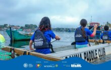 Fotografia horizontal em plano detalhe, tirada de dentro de um barco. Em primeiro plano, a lateral branca do barco de remo é visível, com as palavras "PADDLE CLUB" em preto. Três jovens de costas, vestindo camisetas azuis e coletes salva-vidas pretos, estão sentados na embarcação, remando. Os coletes têm o logo "Passaúna Paddle Club" e "TEAM MIRAGE". Uma jovem de cabelos escuros e colete preto está em posição central, remando com vigor. À esquerda, outra jovem, e à direita, a cabeça de um terceiro remador. No segundo plano à esquerda, uma canoa verde-água flutua. Ao fundo, a água calma e escura do rio. A margem, no horizonte, mostra casas, árvores e uma ponte de estrutura metálica. O céu é totalmente cinza e nublado. A luz é difusa. A cena captura os jovens em plena atividade, praticando o esporte náutico.