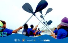 Fotografia horizontal, ângulo de baixo para cima. 7 jovens, de costas, erguem remos pretos com cabos prateados em um barco. Eles usam camisetas azuis e coletes salva-vidas pretos. A ação está focada nos remos cruzados no ar, formando um V invertido central contra o céu branco e claro. À esquerda, um jovem de boné verde e colete laranja segura um remo. No canto direito, uma jovem de gorro roxo, com parte do rosto e ombro visíveis. No centro, 5 jovens de costas erguem seus remos, com rostos voltados para frente, observando o horizonte. Abaixo deles, a água do rio ou mar escura e calma. No fundo, a paisagem é indistinta e nebulosa sob a luz difusa. A atmosfera é de concentração e esforço coletivo. Em sobreposição na base, uma onda azul com os logos de patrocínio.