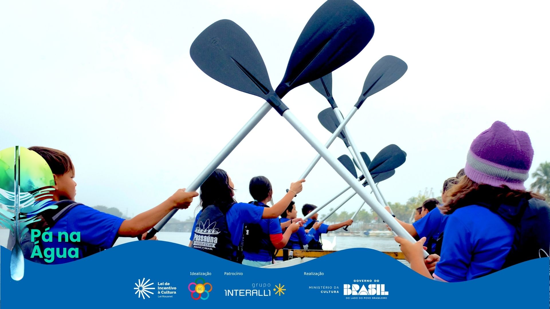 Fotografia horizontal, ângulo de baixo para cima. 7 jovens, de costas, erguem remos pretos com cabos prateados em um barco. Eles usam camisetas azuis e coletes salva-vidas pretos. A ação está focada nos remos cruzados no ar, formando um V invertido central contra o céu branco e claro. À esquerda, um jovem de boné verde e colete laranja segura um remo. No canto direito, uma jovem de gorro roxo, com parte do rosto e ombro visíveis. No centro, 5 jovens de costas erguem seus remos, com rostos voltados para frente, observando o horizonte. Abaixo deles, a água do rio ou mar escura e calma. No fundo, a paisagem é indistinta e nebulosa sob a luz difusa. A atmosfera é de concentração e esforço coletivo. Em sobreposição na base, uma onda azul com os logos de patrocínio.