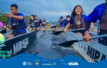 Fotografia horizontal em plano detalhe, tirada de dentro de um barco de remo, focando no centro da embarcação. Cerca de 8 jovens estão visíveis, remando na água escura do rio. Todos vestem camisetas azuis e coletes salva-vidas pretos, com os logos "Team Mirage" e "Passaúna". Em primeiro plano, uma jovem à direita e um jovem à esquerda sorriem enquanto remam vigorosamente. A jovem da direita, de cabelos escuros e longos, tem um sorriso largo e olhos brilhantes. O remo de ambos é preto e largo. As laterais do barco são brancas, com a inscrição "MIRAGE" em preto. A água no centro do barco está agitada pelo movimento dos remos. Em segundo plano, outros jovens remam. O fundo é composto por vegetação densa e um píer, sob um céu de fim de tarde ou nublado, em tons cinza-escuro e azul. A cena captura o esforço e a alegria da atividade em grupo.