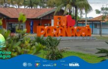 Fotografia horizontal em plano médio de paisagem urbana. No centro, o monumento "EU AMO PARANAGUÁ" em letras de caixa alta, grandes e tridimensionais, na cor laranja vibrante. A palavra "AMO" é substituída por um coração vermelho. O monumento está sobre um canteiro de grama verde, com folhagens e arbustos ao redor. Em primeiro plano, vegetação verde escura em ambos os cantos laterais desfoca a imagem, enquadrando o monumento. Ao fundo, uma construção de alvenaria com telhado de telha de barro marrom-avermelhada e paredes laranjas e bege. Palmeiras e coqueiros estão atrás e à frente do prédio. O céu está cinza-claro, indicando dia nublado. A iluminação é difusa. O monumento é um ponto de referência da cidade. No rodapé, a faixa azul do projeto "Pá na Água" e logotipos de patrocinadores.