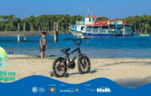 Fotografia horizontal em plano geral na margem de um rio em dia ensolarado. Em primeiro plano, no centro, uma bicicleta infantil preta, com rodinhas laterais e guidão alto, está apoiada na areia clara. A roda dianteira e a coroa do pedal brilham sob a luz forte. No segundo plano, à esquerda, um menino de pele morena clara, cabelo escuro, camisa cinza e short preto, está em pé na água rasa do rio, olhando na direção da bicicleta. Atrás dele, o píer de madeira escuro. No centro e ao fundo, a água azul-escura com pequenas ondas. Uma grande embarcação de dois andares, de casco branco com faixas vermelhas e azuis, está atracada próxima a uma linha de vegetação densa (manguezal). O céu é azul profundo com poucas nuvens. A cena contrasta o brinquedo infantil com o cenário ribeirinho. No rodapé, a faixa azul do projeto "Pá na Água" com logotipos.