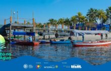 Fotografia horizontal em plano geral, na margem de um rio ou baía em dia de sol. Em primeiro plano, a água escura reflete o sol, com pequenas ondas. A areia clara e larga domina a parte inferior do quadro. Quatro canoas de pesca tradicionais estão atracadas na margem, cobertas por lonas. A primeira canoa à direita é branca com bordas vermelhas. As demais, no centro, têm tons de azul, vermelho e branco, e estão parcialmente cobertas por lonas azuis e cinzas, apoiadas em estruturas de madeira do cais. Um homem de camisa azul está sentado na canoa mais à esquerda. Ao fundo, coqueiros e palmeiras altas criam uma linha verde-escura. À esquerda, uma estrutura de píer rústico e, ao fundo, pessoas caminham na margem. O céu é azul e claro, com luz solar intensa. A cena ilustra o descanso das canoas e o cotidiano do porto.