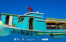 Fotografia horizontal em plano médio e ângulo contrapicado, focando em um barco de dois andares com o nome "Jubai" pintado em preto em seu casco. O barco é predominantemente azul-turquesa vibrante, com detalhes e frisos amarelos ao redor de janelas e convés. Um homem de camisa vermelha e boné está curvado sobre o teto do convés superior, parecendo realizar um reparo ou ajuste. O convés inferior tem uma porta aberta, onde se vê uma pessoa. A embarcação flutua em água calma, sob um céu azul intenso e sem nuvens, que preenche grande parte do quadro. A luz é forte e direta, acentuando o brilho das cores. A cena ilustra o trabalho e a manutenção das embarcações de transporte ou pesca na região. No rodapé, a faixa azul do projeto "Pá na Água" com logotipos de patrocinadores.