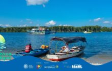 Fotografia horizontal em plano geral na margem de uma baía em dia de sol. Em primeiro plano, duas canoas de pesca tradicionais estão na água rasa, próximas à areia. A canoa da esquerda é azul-clara, e um jovem de camisa vermelha com o número 10 nas costas está sentado. À direita, a canoa é branca e vermelha, coberta por um toldo escuro (sombreiro) e uma pessoa de camisa branca está sentada, com as pernas cruzadas. No segundo plano, a água azul-escura e ondulada. Três barcos maiores estão ao fundo: um barco de dois andares com faixas coloridas (vermelho, branco e azul) está ancorado no centro; à sua direita, um barco de pesca menor e amarelo. O horizonte é dominado por uma densa linha de vegetação verde escura (manguezal). O céu é azul claro com algumas nuvens brancas, e a luz solar é intensa. A cena ilustra o descanso e o transporte no ambiente ribeirinho. No rodapé, a faixa azul do projeto "Pá na Água" com logotipos.