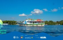 Fotografia horizontal em plano médio de um barco de transporte ou turismo flutuando em água azul-escura. A embarcação é de dois andares, de casco branco, e pintada com listras horizontais vibrantes em azul, verde, vermelho e amarelo. O convés superior exibe um toldo branco e possui equipamentos de navegação. Uma boia salva-vidas vermelha com detalhes em branco está pendurada na lateral. O barco está no centro da imagem, e a água apresenta pequenas ondulações. O fundo é delineado por uma densa linha de vegetação verde (manguezal ou floresta de restinga) que acompanha o horizonte. O céu é azul intenso, com duas nuvens brancas e fofas acima da embarcação. A luz solar é forte, realçando o contraste das cores. Um pequeno barco branco é visto flutuando à esquerda, perto da vegetação. A cena remete ao transporte e ao turismo aquático sob clima tropical. No rodapé, a faixa azul do projeto "Pá na Água" com logotipos.