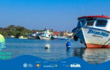 Fotografia horizontal em plano médio na baía, sob céu azul. No primeiro plano, à direita, a proa de um barco de pesca ou turismo, chamado "Brisa Mar Adventure", ocupa a maior parte da lateral. O barco tem casco branco, detalhes em azul e a borda superior em vermelho, destacando-se na diagonal. Uma boia azul e uma boia retangular cinza flutuam na água escura, presas ao barco por cabos. No segundo plano, uma fileira de embarcações de tamanhos variados está atracada próximo à margem. Estas incluem barcos de dois andares, brancos com detalhes coloridos (azul e verde), e pequenos barcos de pesca. A margem é densamente arborizada com vegetação escura. O céu azul intenso, com algumas nuvens brancas, reflete-se na água calma, que apresenta leves ondulações. A cena remete a um porto ativo e à diversidade de embarcações da região. No rodapé, a faixa azul do projeto "Pá na Água" com logotipos.