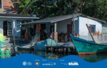 Fotografia horizontal em plano médio de uma moradia ribeirinha rústica. A casa, branca e simples, tem um telhado de telha de amianto e paredes de alvenaria, com trechos de tijolo aparente. Roupas estão penduradas sob a varanda central. No centro, uma área de píer ou deck de madeira precário está sobre a água. Redes de pesca (tarrafas) estão penduradas secando próximo à varanda. Uma canoa de madeira, com o casco verde-azulado, está atracada à direita, em primeiro plano, na água calma e escura. A lateral esquerda mostra uma casa de alvenaria branca com porta e janelas grandes. Acima e atrás da casa, uma densa vegetação tropical (bananeiras e folhagens escuras) cria o fundo. A iluminação é de sol forte, com sombras nítidas. A cena retrata o cotidiano de pesca e a arquitetura simples e funcional da vida ribeirinha. No rodapé, a faixa azul do projeto "Pá na Água" com logotipos.