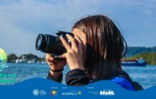 Fotografia horizontal, plano médio. Uma jovem, perfil à direita, segura uma câmera DSLR preta, mirando o visor com o olho esquerdo. Seu rosto e olhos estão ocultos pela câmera. O cabelo escuro, molhado e lustroso, cai sobre os ombros. veste uma blusa preta de manga longa. A jovem é o foco nítido e ocupa o centro da imagem. O fundo é o rio ou mar em azul escuro, totalmente desfocado (bokeh). Na paisagem em segundo plano, aparecem pontos brancos de pequenos barcos atracados à esquerda e uma grande embarcação de casco azul-escuro à direita. A vegetação densa e escura da mata ribeirinha preenche a linha do horizonte. O céu claro reflete na água. Na parte inferior, há uma faixa azul ondulada com o logo do projeto e os créditos.