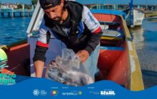 Fotografia em plano médio, tirada de cima para baixo, focando em um homem trabalhando dentro de uma canoa de pesca. O homem, de pele morena, barba e boné preto, veste um casaco esportivo preto, branco e vermelho e calça jeans clara. Ele está curvado dentro da canoa, usando as duas mãos para segurar um saco plástico transparente, de onde retira peixes grandes e prateados. O interior da canoa é de um vermelho-alaranjado vibrante, com a borda externa amarela. A canoa flutua em água rasa e clara. Ao fundo, uma canoa de cor bege está na margem e, além dela, uma estrutura de píer de madeira rústica e a linha de vegetação ribeirinha. O pescador está concentrado na atividade. O sol brilha no rosto do homem e na água, refletindo o céu azul ao fundo. A cena representa o momento do desembarque da pesca. No rodapé, a faixa azul do projeto "Pá na Água" com logotipos.