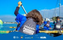 Fotografia em plano médio, tirada de um ângulo ligeiramente baixo, capturando o esporte de canoagem em equipe. Em primeiro plano, uma jovem de cabelos castanhos escuros e casaco preto e azul está de costas, com os braços erguidos, segurando um remo prateado em posição de remada. No seu colete salva-vidas preto, lê-se "Passaúna Paddle Club - Paraná Brasil" em branco. À frente e ao lado dela, outros jovens, todos vestindo coletes pretos e camisas azuis, remam em sincronia. A canoa, de borda alta, tem listras pretas na parte superior. O grupo está em movimento sobre a água azul escura. Ao fundo, a luz do sol brilha na água e revela mastros e velas de veleiros brancos atracados, além da linha de árvores e casas da margem. A cena retrata o entusiasmo e a coordenação do esporte aquático em equipe. No rodapé, a faixa azul do projeto "Pá na Água" com logotipos.