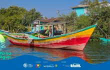 Fotografia horizontal em plano médio de uma canoa de pesca de madeira ancorada em água rasa. A canoa, no centro da imagem, é pintada com cores fortes: uma faixa vermelha escura na parte inferior, e uma faixa amarela vibrante na parte superior, delimitada por uma linha verde-água. A proa, à direita, é alta e inclinada, destacando-se na diagonal. A água é escura e calma, refletindo ligeiramente o casco. Em primeiro plano, o barco flutua próximo a uma densa vegetação verde escura (manguezal), que cobre a maior parte da margem. Ao fundo, entre a folhagem, notam-se construções rústicas coloridas: uma casa à direita é azul-turquesa, e outras têm telhados de zinco aparente. Redes de pesca (tarrafas) estão penduradas entre as construções. A cena é um registro da arte e do cotidiano ribeirinho em um dia de sol. No rodapé, a faixa azul do projeto "Pá na Água" com logotipos.