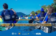 Fotografia em plano médio, tirada de um ângulo ligeiramente baixo, capturando uma equipe de jovens remando em uma canoa de competição. Em primeiro plano, a imagem é dominada pelas costas e ombros dos remadores. Eles vestem camisas azuis e coletes salva-vidas azuis ou pretos, com os logotipos brancos "Passaúna Paddle Club" ou "Team Mirage". Os remadores estão alinhados, com remos pretos imersos na água azul escura, criando pequenas ondulações. A canoa, branca e preta, tem tábuas de madeira marrom clara atravessando o topo. Ao fundo, uma segunda canoa com mais remadores segue o grupo principal. O horizonte é composto por morros de mata atlântica densa, em tons de verde escuro e azulados, e sob um céu claro. A cena é dinâmica e ilustra a coordenação e o esforço do esporte de canoagem em equipe. No rodapé, a faixa azul do projeto "Pá na Água" com logotipos.