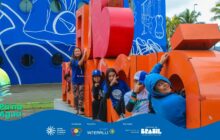 Fotografia em plano médio de um grupo de seis crianças e adolescentes interagindo com um grande letreiro de letras em 3D, pintado de laranja e vermelho. O letreiro é parte de uma frase turística, mas as crianças se apoiam ou se sentam nas letras. Os participantes vestem camisas azuis do projeto e bonés. Em primeiro plano, uma menina à esquerda sorri, apoiada na lateral de uma letra vermelha. À direita, um menino com capuz azul sorri, inclinado para fora de uma letra laranja. As outras quatro crianças estão no centro, posando para a câmera. O fundo é dominado por uma grande parede de alvenaria pintada em azul vibrante, com desenhos em branco, e por troncos de palmeiras. Ao fundo, no horizonte, morros arborizados em tons azulados. A cena transmite a alegria e a energia dos jovens no espaço urbano e turístico da cidade. No rodapé, a faixa azul do projeto "Pá na Água" com logotipos.