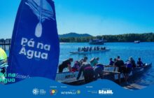 Fotografia em plano aberto, capturando uma cena de esporte náutico em um dia ensolarado. Em primeiro plano, à esquerda, uma grande vela ou bandeira vertical, pintada de azul profundo, exibe o logotipo e o nome "Pá na Água" em letras brancas, ocupando a maior parte da lateral esquerda e do centro inferior da imagem. No centro, uma canoa de competição (outrigger ou barco-dragão) é retirada da água por um grupo de remadores (homens e mulheres), todos de costas, vestidos com roupas de esporte e bonés. O casco escuro da canoa exibe a escrita "Team Mirage" e "Passaúna" nas laterais. No meio-plano, outra canoa de competição flutua, com remadores sentados. Ao fundo, na água azul escura, um barco de passageiros branco e azul (traineira) navega, e a margem é delimitada por uma linha densa de mata. O céu é de um azul vívido, com o sol brilhando intensamente. A cena celebra o esforço coletivo e o cenário natural da baía. No rodapé, a faixa azul do projeto "Pá na Água" com logotipos.