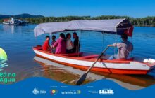 Fotografia em plano médio, capturando um momento de travessia em uma baía ou rio largo. No centro, uma canoa de pesca de madeira, com o casco vermelho e branco, tem um toldo de lona branca que protege os passageiros do sol. Dentro da canoa, quatro pessoas (duas adultas e duas crianças) estão sentadas de costas para a proa, conversando e olhando para trás. Na popa (parte traseira), um homem de boné escuro, camisa cinza e calça jeans, sentado de lado, segura um remo que usa para impulsionar e guiar a embarcação. A água azul escura é calma, e o reflexo do sol brilha na superfície. No plano de fundo, à esquerda, uma balsa ou barco de passageiros branco e azul navega, e a margem oposta é densamente coberta pela vegetação verde escura da mata atlântica. O céu azul intenso sugere um dia claro e ensolarado. A cena é um registro do transporte e turismo em comunidades ribeirinhas. No rodapé, a faixa azul do projeto "Pá na Água" com logotipos.