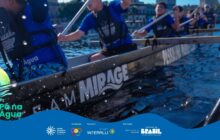 Fotografia em plano fechado, tirada dentro de uma canoa de competição (Outrigger Canoe ou Barco-dragão), capturando a ação de um grupo de remadores em um dia ensolarado. A cena é dinâmica e centrada na água: respingos de água cobrem as laterais da canoa preta e escura, onde se lê a inscrição "TEAM MIRAGE" em letras brancas grandes. Quatro jovens remadores estão visíveis: dois à esquerda e dois à direita (o braço de um quinto remador está no canto inferior direito). Eles vestem camisas azuis e coletes salva-vidas pretos, com os logos do projeto. Seus braços estão levantados e engajados no movimento de remar, com os remos fora de vista no momento da foto, exceto pelo cabo na lateral. A expressão facial de esforço, combinada com a luz forte e os respingos, confere à imagem uma sensação de velocidade e intensidade. Ao fundo, estruturas urbanas e árvores estão desfocadas. No rodapé, a faixa azul do projeto "Pá na Água" com logotipos.