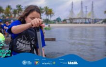 Fotografia em plano fechado, com profundidade de campo rasa, focada em uma jovem participante do projeto. Em primeiro plano, no centro, uma menina (criança ou pré-adolescente), com longos cabelos castanhos escuros, está ajoelhada ou inclinada, segurando firmemente o cabo de um remo preto. Ela veste uma camisa azul e um colete salva-vidas preto com o logotipo branco "Passaúna Paddle Club". Seus olhos estão voltados para a areia molhada e a água rasa onde ela pisa. A luz do dia é difusa, mas ilumina seu rosto e a lateral do colete. Atrás dela, desfocada, a praia se estende até a água. No plano de fundo, estruturas brancas de telhado cônico (semelhantes a tendas de porto) se destacam contra o céu claro. A cena é emoldurada por coqueiros, e à esquerda, outras pessoas, também vestidas de azul, estão visíveis em segundo plano, sugerindo o início de uma atividade. A foto capta o foco e a concentração da jovem atleta. No rodapé, a faixa azul do projeto "Pá na Água" com logotipos.